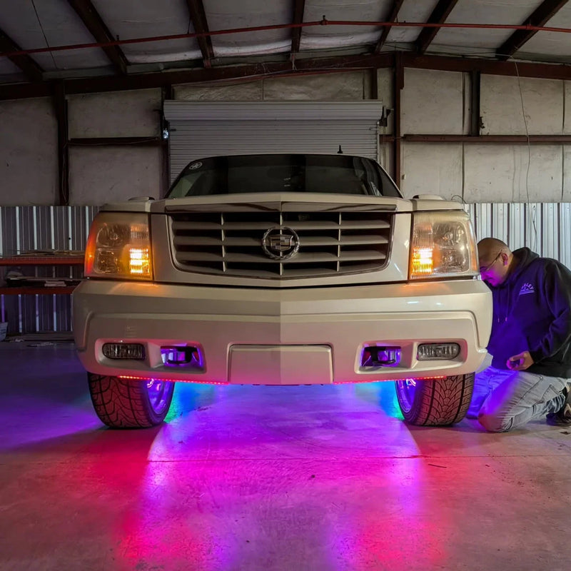 Silver Cadillac Escalade in a shop with amber headlights and pink and blue underglow lights while an installer kneels beside it